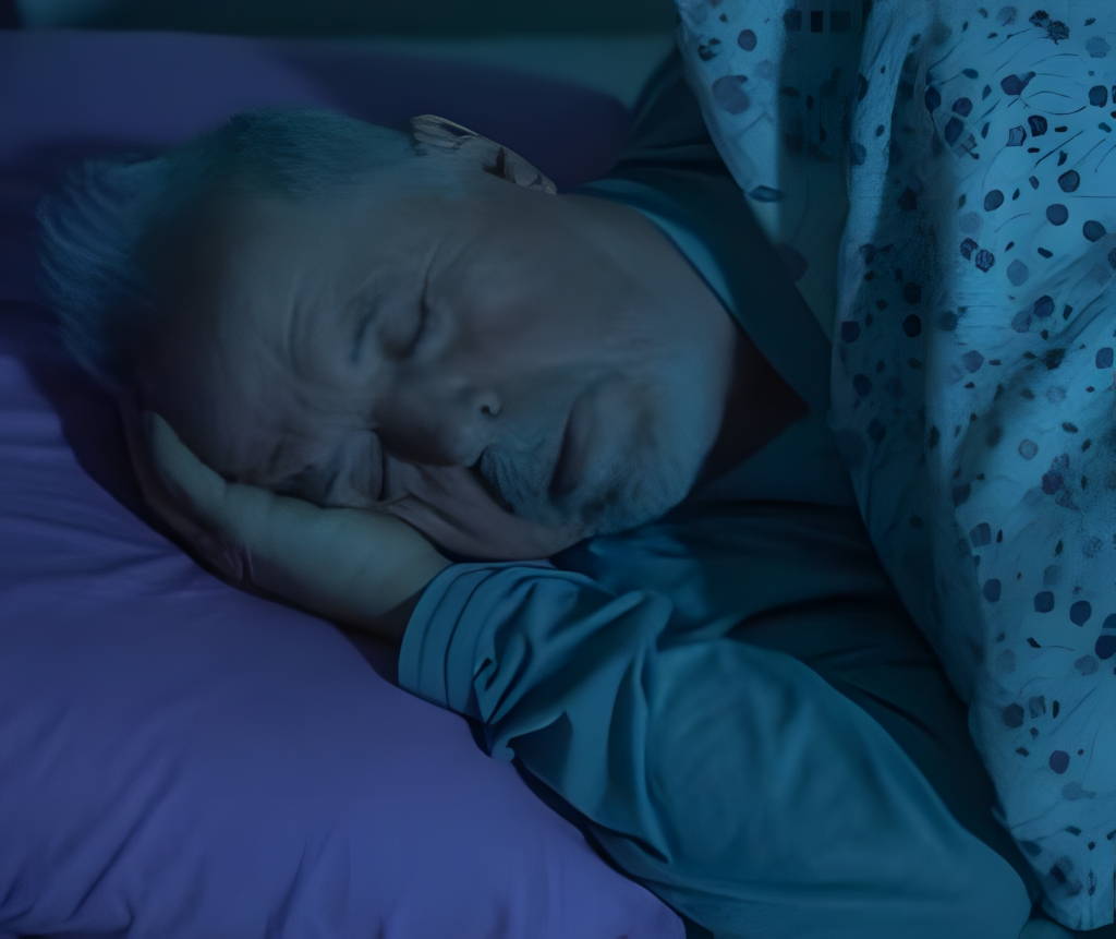 A man with short gray hair and a relaxed expression is comfortably sleeping on a purple pillow under a patterned blanket, illuminated by soft, dim blue lighting. Resting his head on his hand, he appears peacefully tucked in bed at home. | Bed For Seniors