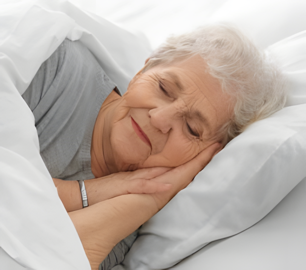 An elderly woman with short white hair is peacefully sleeping in her bed at home, resting her head on a white pillow and lying under a cozy white blanket. She has her hands tucked under her cheek and is wearing a light gray shirt. | Bed For Seniors