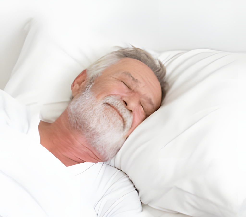 An older man with white hair and a beard is lying in bed at home, smiling with his eyes closed. He is resting his head on a white pillow and is covered with white sheets. The scene suggests peaceful sleep or relaxation. | Bed For Seniors