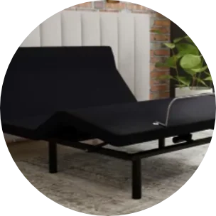A modern adjustable bed for seniors with a black frame and black mattress is set in a bedroom with a gray carpet. There is a potted plant and a brick wall with white paneling in the background. | Bed For Seniors